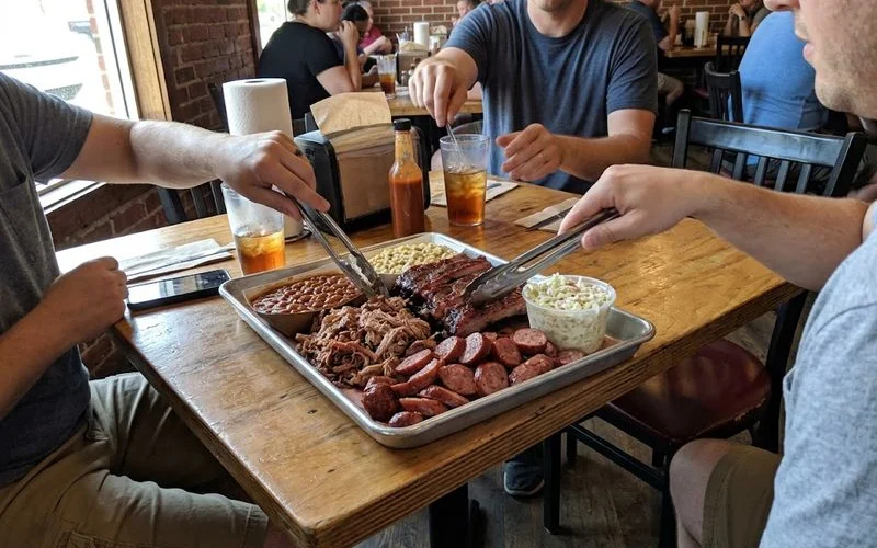 A family-style BBQ platter piled with brisket, ribs, and sides meant to be shared across a table, next to a stack of plates